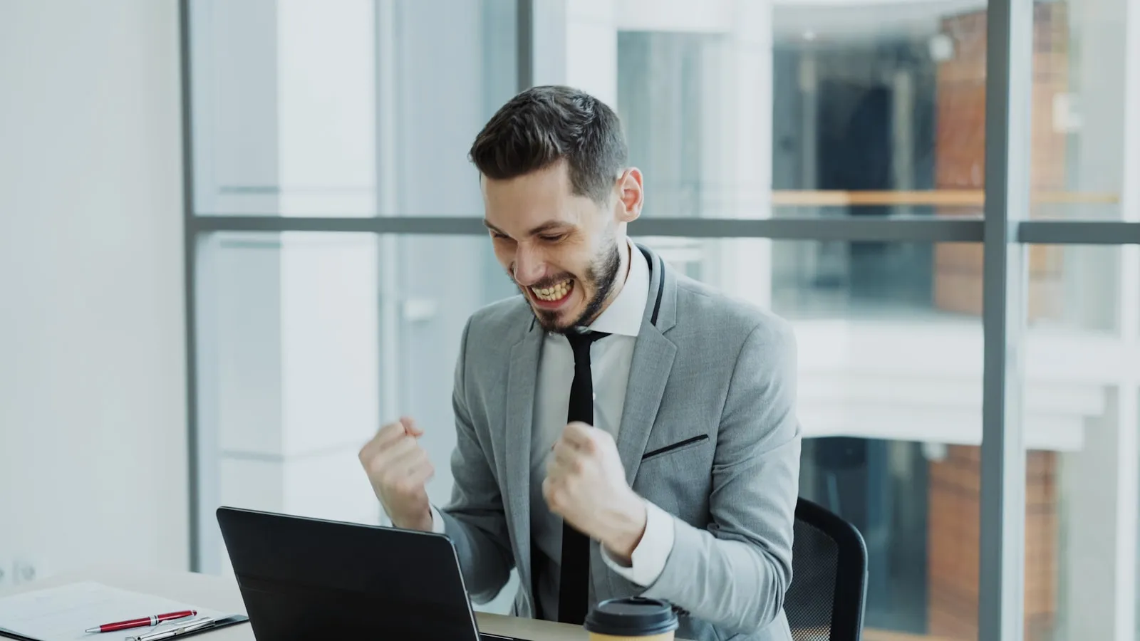 Happy man celebrating success with a fist pump and smile while working at a laptop in a modern office.