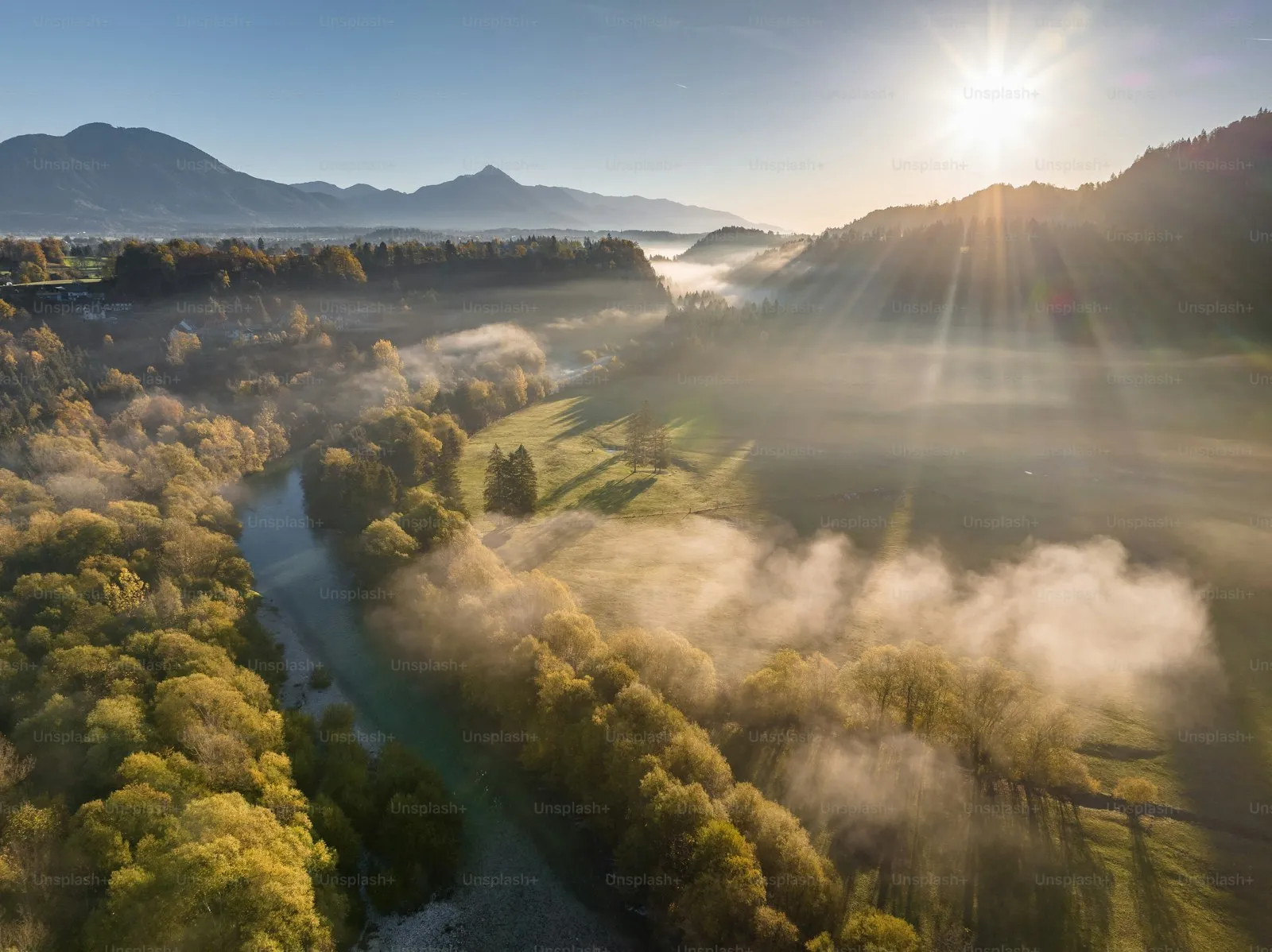 Aerial view of lush green forests, a winding river, and misty mountains under bright sunlight in a scenic natural landscape.