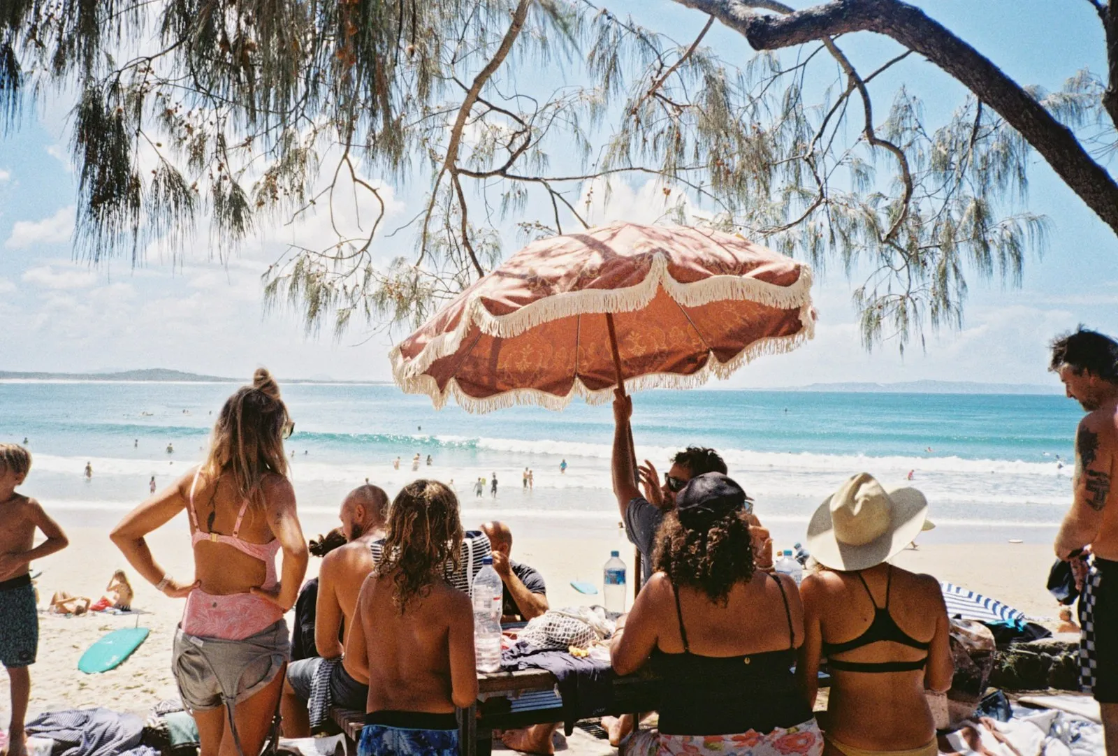 A diverse group of people enjoying a sunny day on the beach under a large umbrella, with ocean and sky in the background.