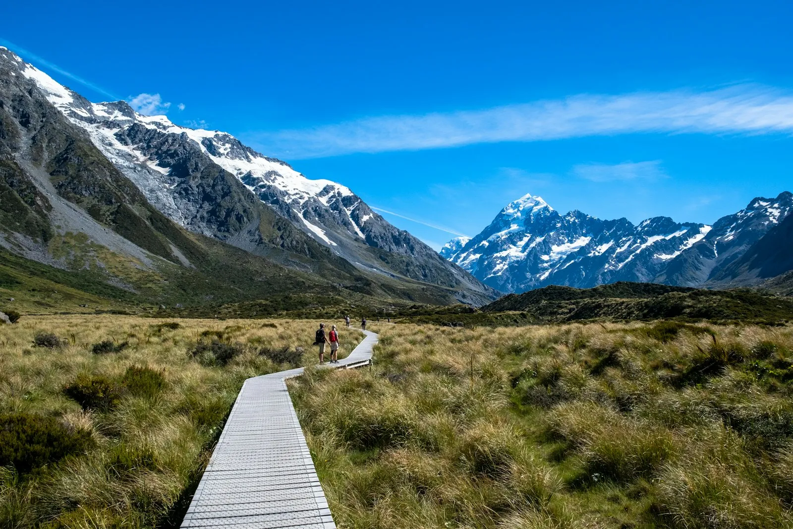 Vast mountain landscape in New Zealand with hikers walking along a wooden trail under a bright blue sky.