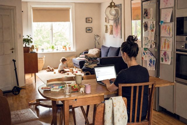 A woman typing a first message at a cluttered family dining table, a toddler playing on the rug behind her
