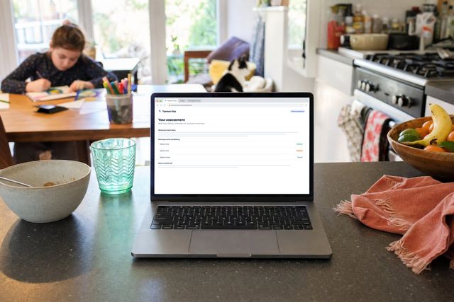 A laptop on a kitchen worktop showing a Tasman Visa written assessment, a child colouring at the table behind