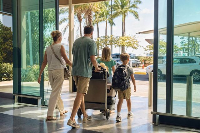 A family walking out through airport arrivals into the sun with a loaded luggage trolley